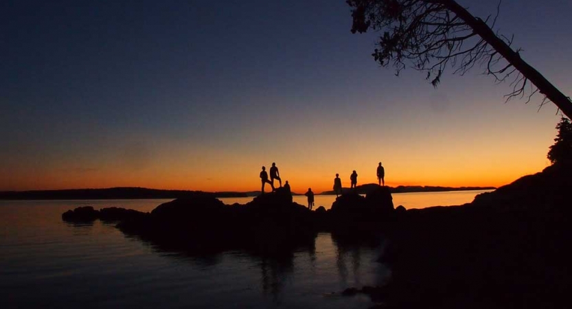 the silhouette of a group of people standing on rocks jutting out of a body of water is illuminated against the sunset.